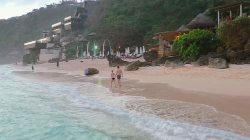 aerial coastline of young couple walking on a white sand beach at Uluwatu cliffs in Bali at sunset