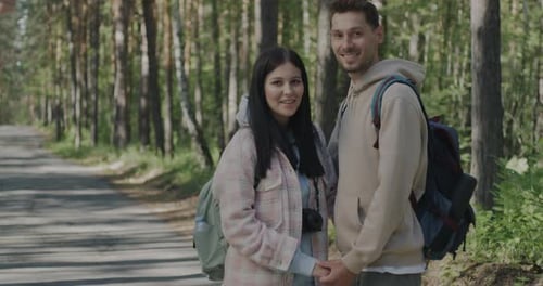 Joyful Young People Couple with Backpacks Standing in Forest Holding Hands Smiling Looking at Camera