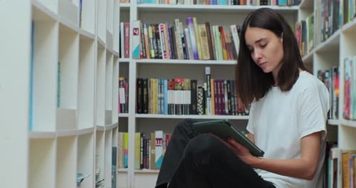 Caucasian Girl Sitting on the Floor Near Shelves with Book in the Library Uses Digital Tablet