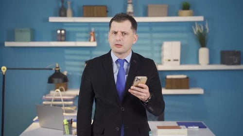 A businessman in a suit receives a message on his phone in his office and gets angry at the bad news