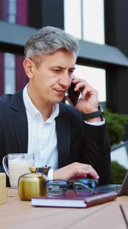 Middleaged Businessman Working on Laptop at an Outdoor Cafe While Having a Business Call on Phone
