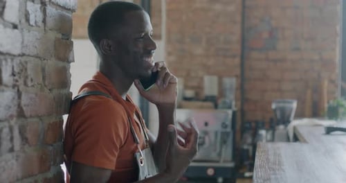 Young African American Man Barista Talking on Mobile Phone Standing at Counter in Cafe