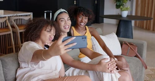 Amigas afro-americanas no sofá, mulher deixada estendendo o smartphone verificando selfies, reagindo
