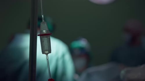 Closeup Medical Dropper in Darkness in Operating Room with Blurred Surgeon and Nurses Performing