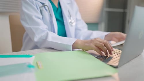 Female doctor in lab coat typing on laptop while working at table in medical office