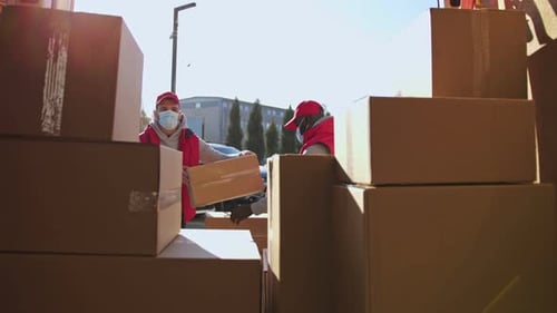 Afroamerikanische und kaukasische Kuriere mit Gesichtsmasken transportieren Paketkästen von einem Trolley in einen Van