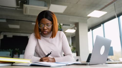 Woman Writes in Notebook at Desk with Laptop