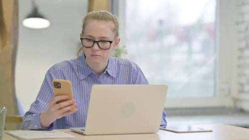 Woman Using Phone and Laptop at Desk