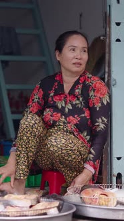Barefoot Mature Asian Woman Seated Next to a Fan Waits for Customers at a Stall in the Local Market