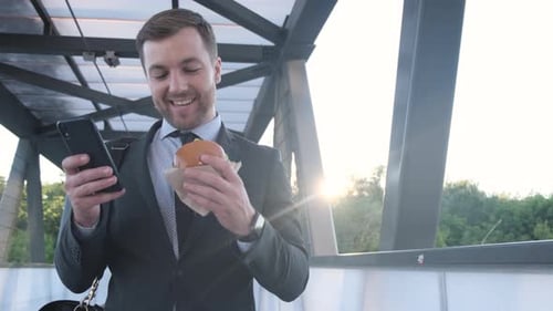 Young Adult Man Eating Burger and Using Phone