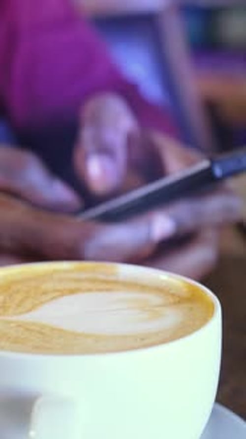 Gros plan d'une tasse à café avec latte art et mains floues à l'aide d'un smartphone dans un cadre de café confortable