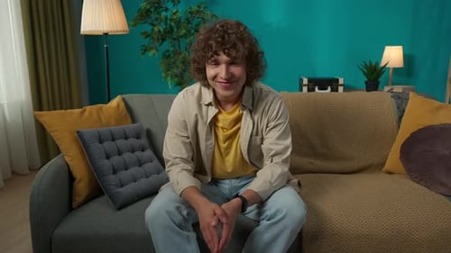 Junger Mann in Freizeitkleidung sitzt auf dem Sofa im Wohnzimmer und schaut in die Kamera und
