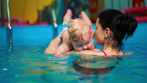 Rear view of a woman holding a baby in the swimming pool. Coach is teaching an infant to swim.