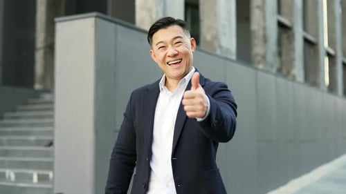 Smiling asian businessman in formal suit showing thumb up looking at camera while standing on street