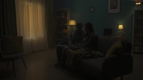 A Couple is Sitting on a Sofa in the Dark in the Room A Man Listens to a Woman Holds Her Hand Hugs