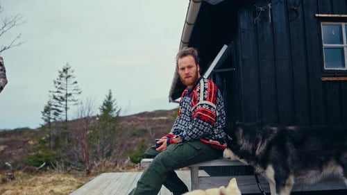 Norwegian Guy With Alaskan Malamute Dogs Resting Outside The Mountain Lodge. Static Shot