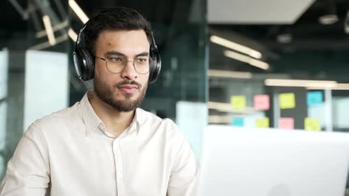 Businessman in wireless headphones works on a laptop sitting at a workplace in business office.