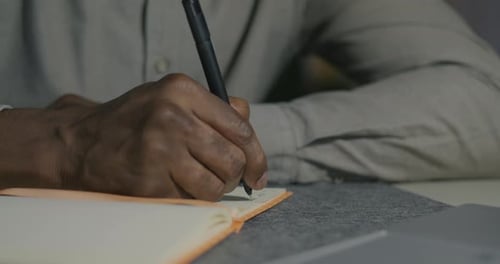 Closeup of Male Hands Writing Information in Notebook While African American Man Working at Desk at