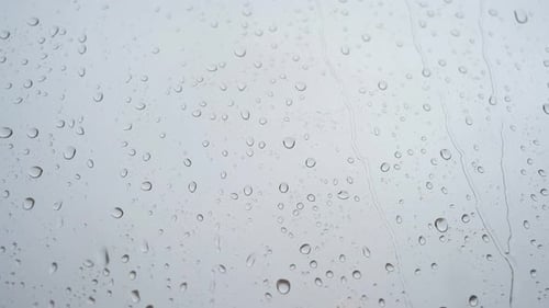 Durch ein Fensterglas ist eine Nahaufnahme schwerer Regentropfen in Zeitlupe zu sehen.