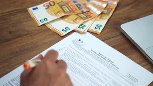 Businessman Fills Out W9 Form with Pen Setting Workplace with Laptop and Several Documents