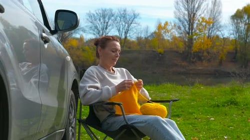 Woman Knitting Outdoors by Car on Autumn Day