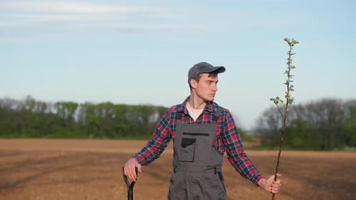A Farmer with a Shovel and a Seedling Stands in a Field