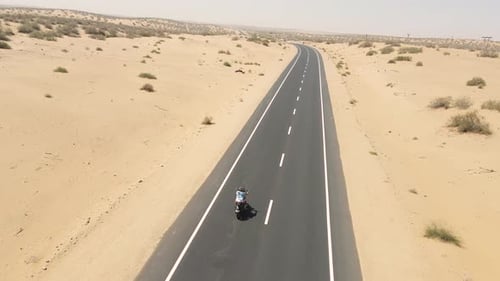 Aerial drone shot of a bike rider navigating curves on a remote desert road surrounded by sand.