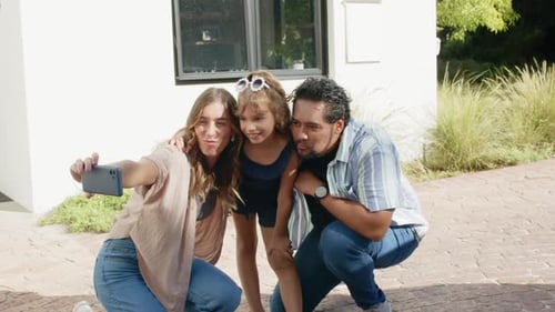 Família diversificada com criança posando na varanda enquanto mulher se agacha, estendendo o smartphone para selfie