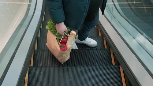 Hombre en una escalera mecánica que sostiene una bolsa de papel ecológica con comida dietética vegetariana en un centro comercial sostenible