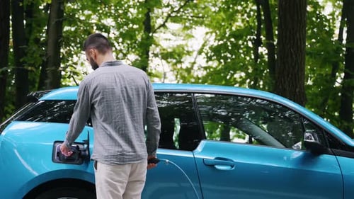 Young man charging electric car outdoor at green forest. Save ecology alternative energy sustainable