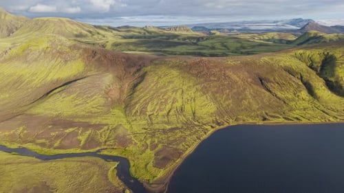 Moss-covered volcanic hills and winding river meet a deep blue lake under soft clouds