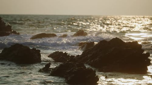 Sunset Glints on Ocean Waves Crashing Against Jagged Rocks at Beach Frothy Sea Foam Bubbles in