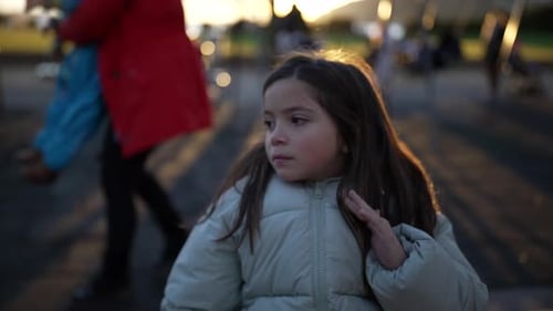 One little girl sitting at park bench during sunset time wearing winter coat and adjusting hair