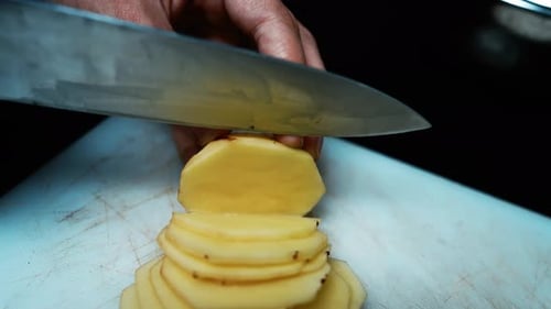 Close-up of hands slicing a peeled potato with a knife on a cutting board