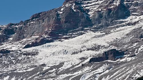 Snow-covered rocky mountain cliffs with exposed strata and glacial ice under a clear blue sky.