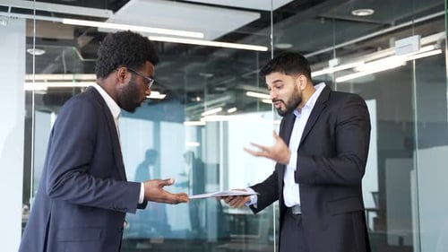 Two angry businessmen standing in modern office arguing over documents on clipboard.