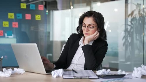 Gelangweilte Büroangestellte sitzt mit Laptop und zerknittertem Papier am Schreibtisch und sieht müde aus.
Frau