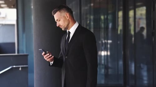 Smiling business man dressed in suit talking by smartphone while standing outside a glass building