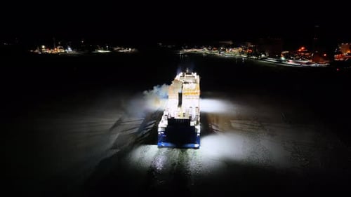 Hybrid roro ship moving through ice covered sea at night. Harbour lights ahead. Aerial stern view.