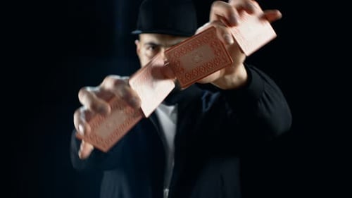 Professional Street Magician in a Cap Performs Sleight of Hand Card Tricks. Background is Black.