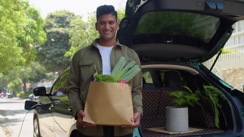 Retrato de hombre con bolsa de comestibles cerca de la carga de un coche eléctrico con cable