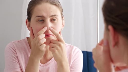 Young woman looking in mirror and squeezing pimples on her nose