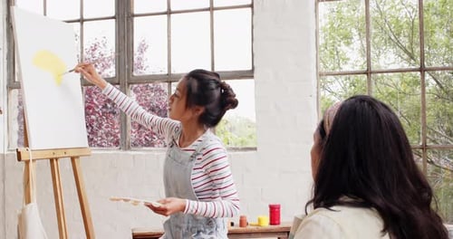 Multiracial young woman painting on canvas while friend observes in bright art studio, at home