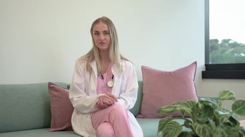 Erwachsene Krankenschwester hört Stimme aus dem Off, die sich zur Kamera wendet und auf einem blaugrünen Sofa in rosafarbenem Peeling lächelt