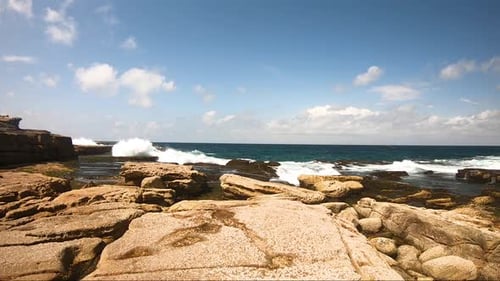 Kleine Wellen krachen über Felsen an einem Strand mit blauem Himmel und Wolken im Hintergrund.