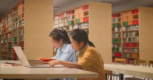 Étudiantes avec un casque lisant des livres tout en étant assise sur la table avec un ordinateur portable dans la bibliothèque