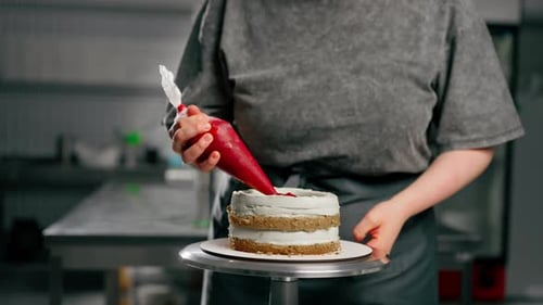 Close Up Female Baker in a Professional Kitchen Puts Red Cream From Pastry Bag Onto a Sponge Cake
