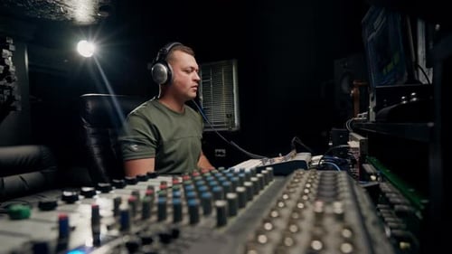 a focused male sound engineer wearing headphones sits at a mixing desk recording new track