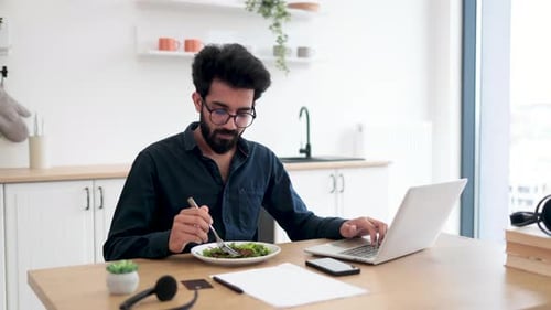 Young Adult Eating Salad While Working on Laptop