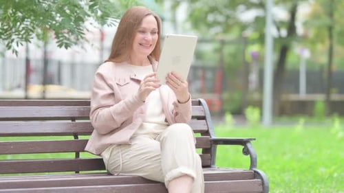 Senior Old Woman Doing Video Chat on Tablet while Sitting Outdoor on a Bench